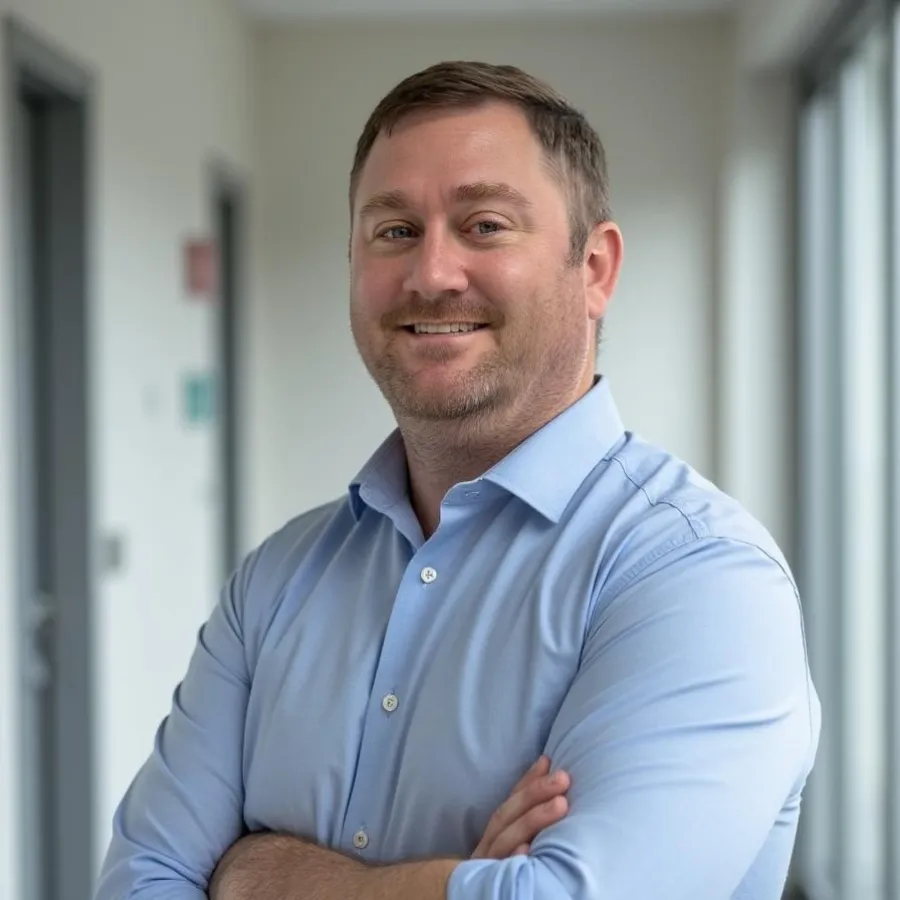 Smiling man with crossed arms wearing light blue shirt in a bright modern office corridor