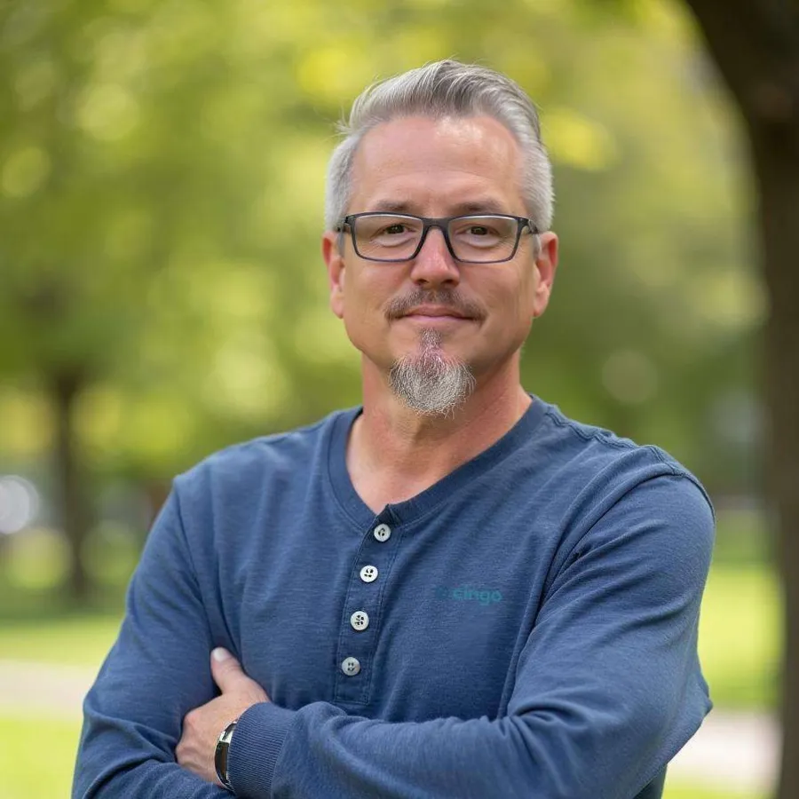 Mature man with gray hair and beard wearing glasses and blue henley shirt standing outdoors with arms crossed.