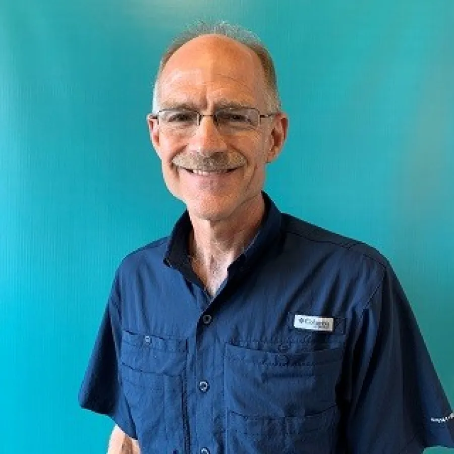 Smiling older man with glasses wearing a navy blue Columbia shirt against a teal background