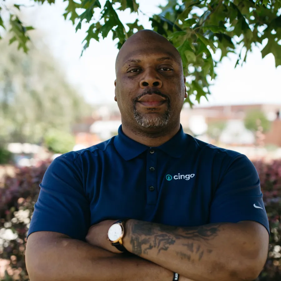 Man with crossed arms wearing a navy blue Cingo polo shirt standing outdoors under tree branches.