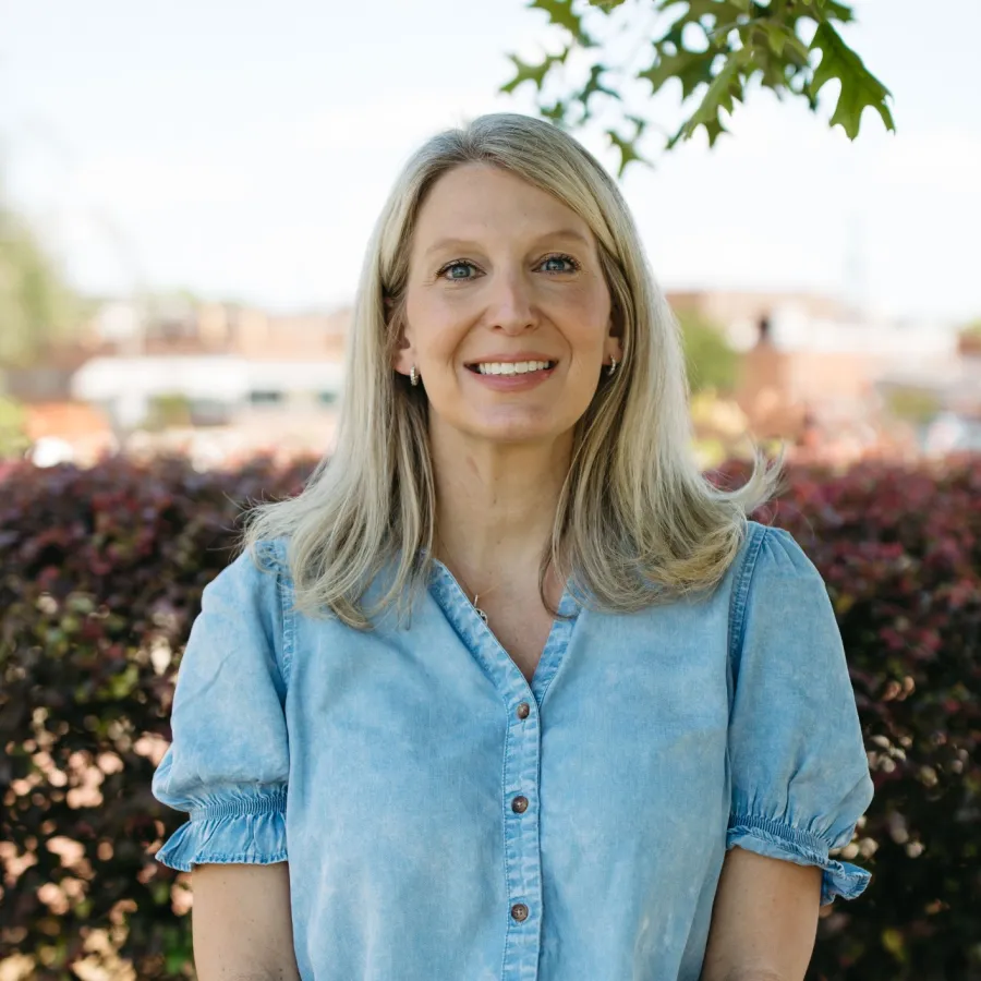 Smiling blonde woman wearing a light blue blouse and white pants standing outdoors with green foliage background