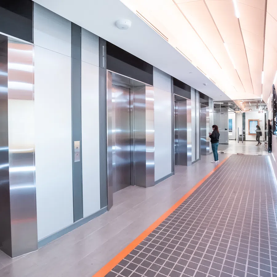 Modern elevator lobby with stainless steel doors, tiled floor corridor, and two people waiting near elevators.