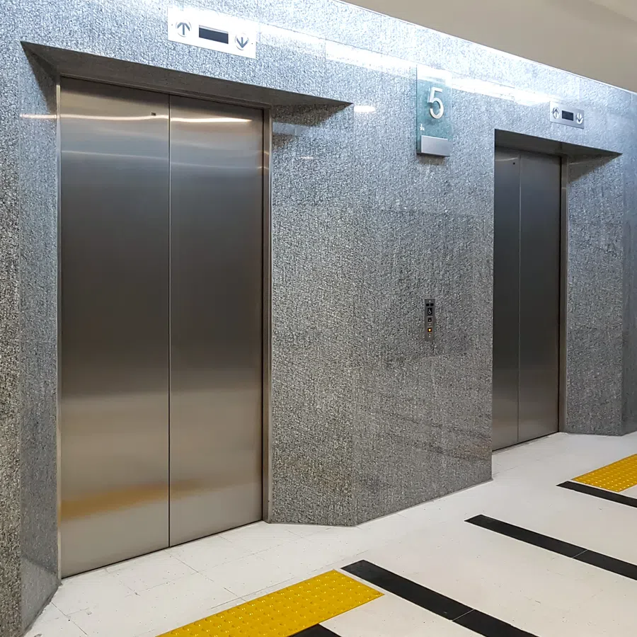 Two closed stainless steel elevators in a modern building hallway with gray walls and floor markings.