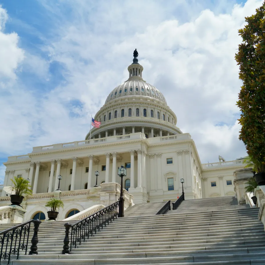 Wide shot of the U.S. Capitol building with stairs, columns, and an American flag against a partly cloudy sky.