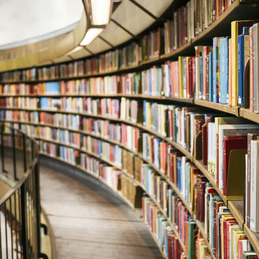 Curved library shelves filled with colorful books lining a wooden walkway with black metal railing and overhead lighting