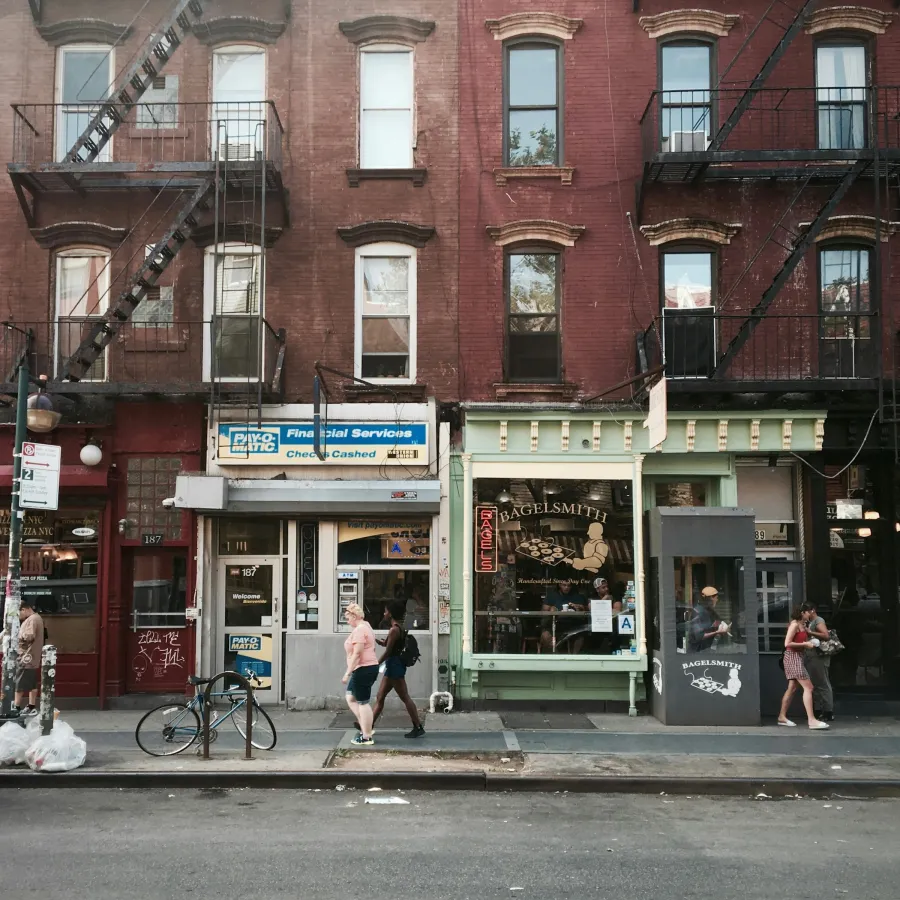 Street view of urban shops with fire escapes, pedestrians, bagel shop, financial services, and parked cars