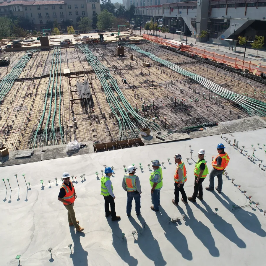 Construction workers in safety gear inspect foundation work with exposed wiring and rebar on a sunny day.