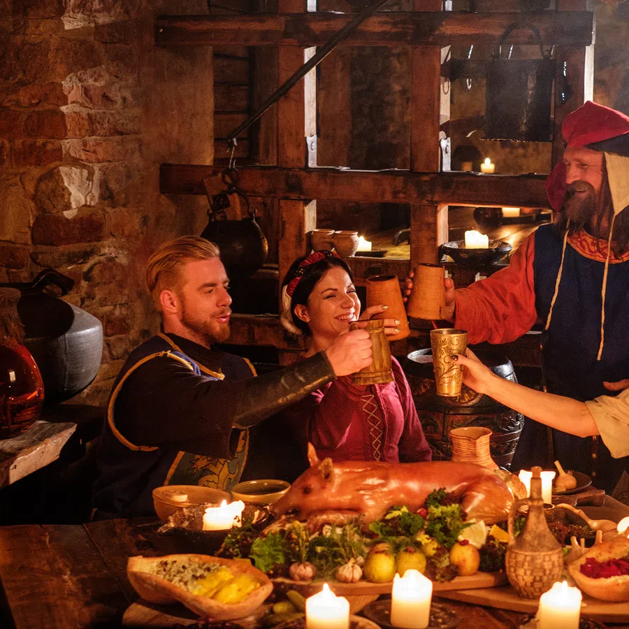 Four people in medieval costumes toasting with mugs around a candlelit feast with a roasted pig and rustic dishes.