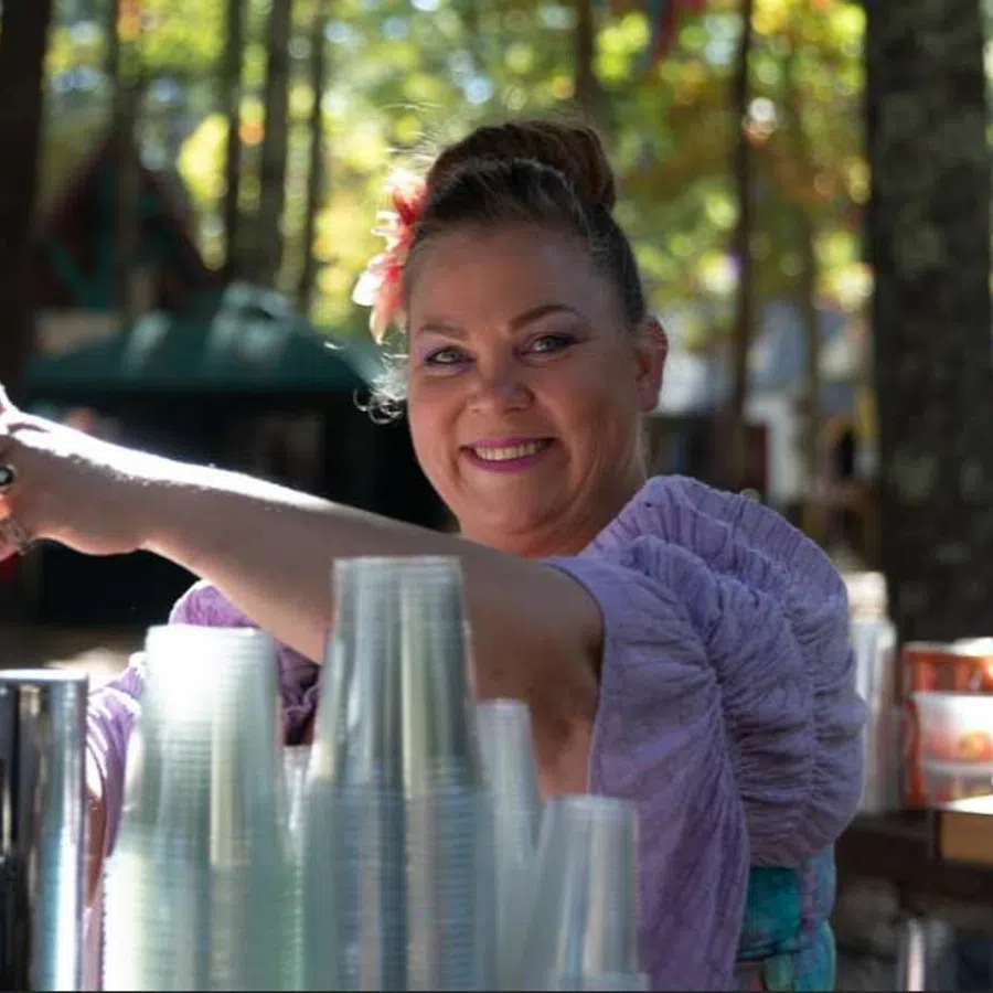 Smiling woman serving drinks outdoors with a flower in her hair and clear plastic cups stacked nearby.