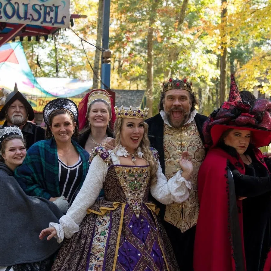 Group of people dressed in colorful medieval costumes posing happily in front of a carousel in autumn.