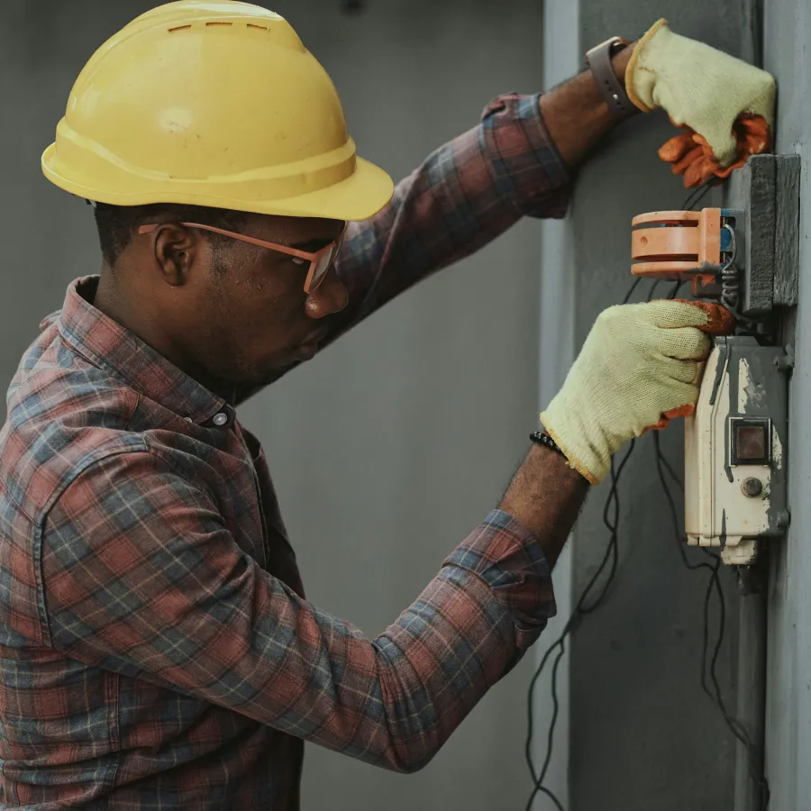 Electrician wearing safety helmet and gloves working on electrical wiring on a gray wall.