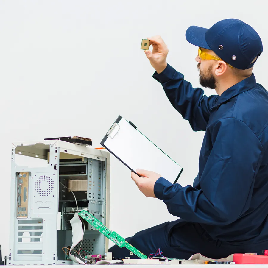 Technician in blue uniform inspecting computer processor while holding clipboard beside open desktop PC.