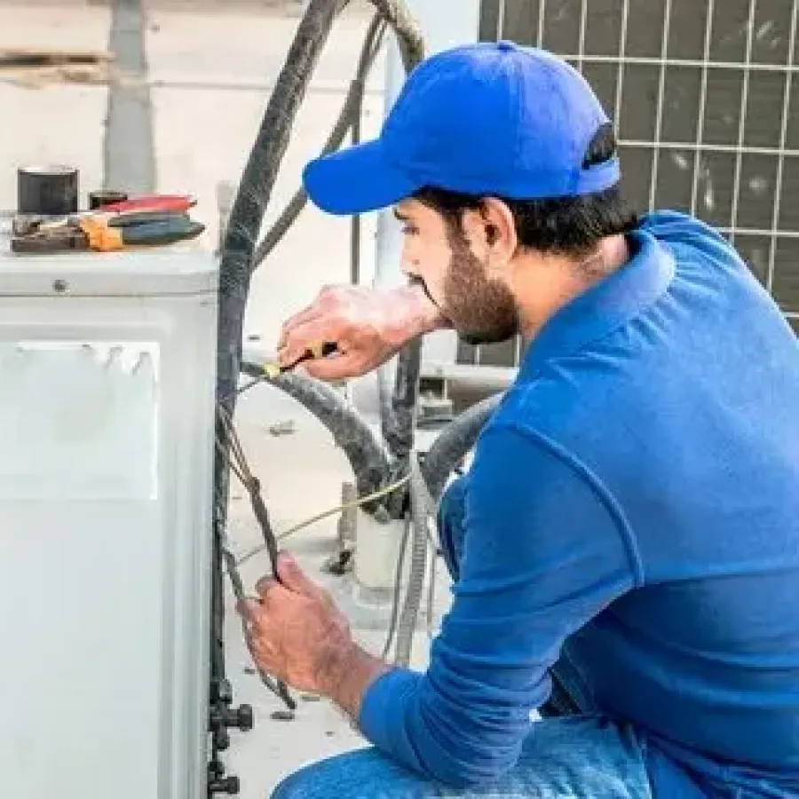 Technician in blue uniform repairing or installing an outdoor air conditioning unit with tools on top.