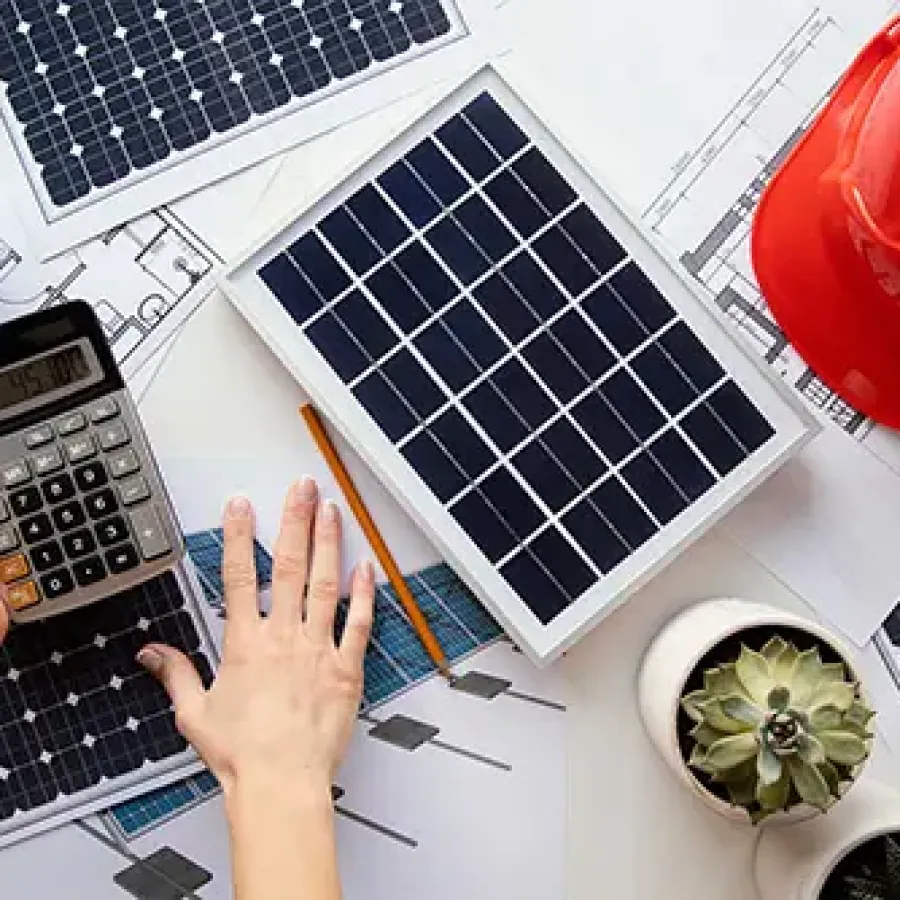 Hands using a calculator on solar panel blueprints with small solar panels, a red helmet, and plants on a table.