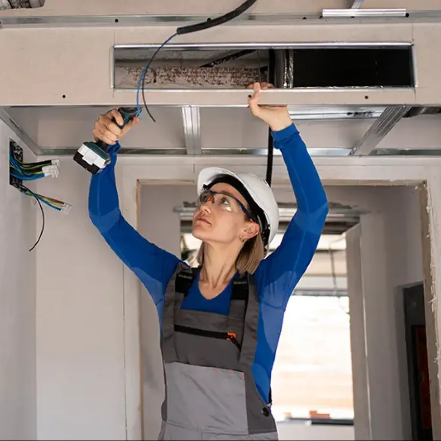 Female electrician wearing safety gear working on ceiling electrical wiring with power drill in unfinished room.