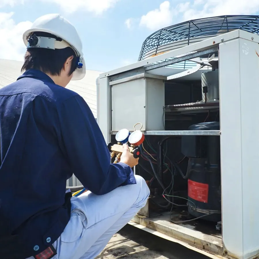 Technician in hard hat inspecting and repairing commercial HVAC unit outdoors under blue sky.