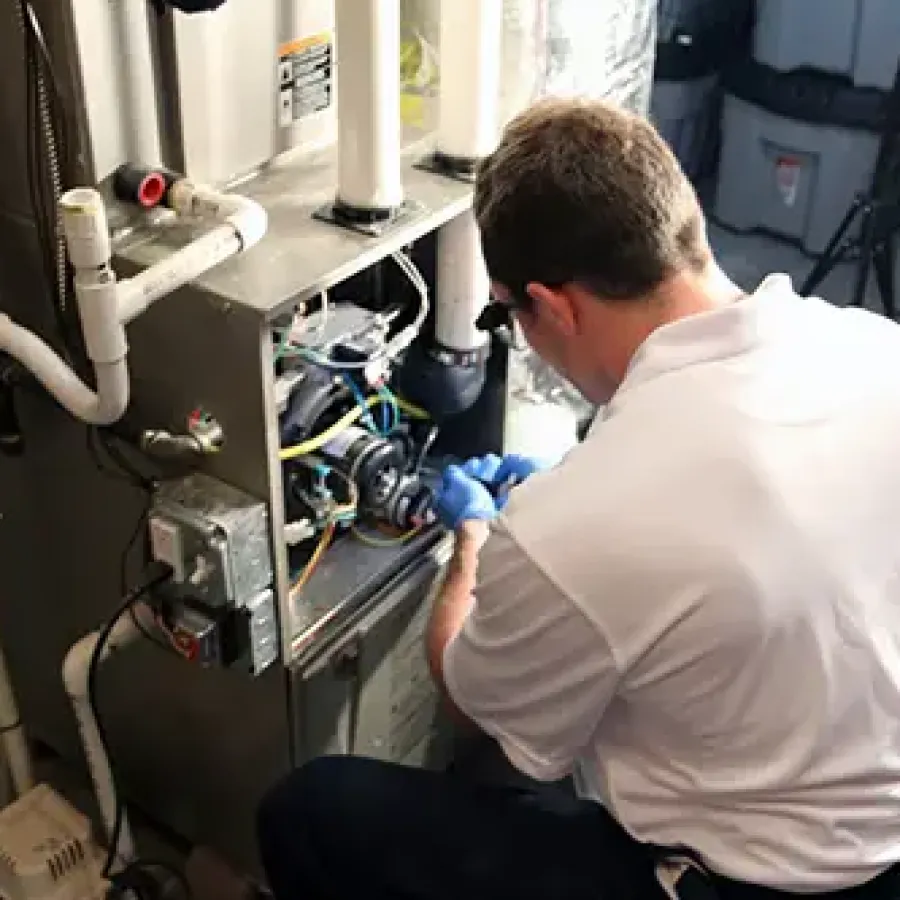 Technician in white shirt repairing a home furnace with blue gloves in a basement setting.