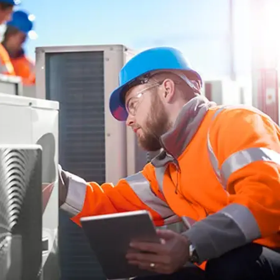 Technician in orange safety gear and blue helmet inspecting HVAC unit outdoors with tablet in hand on a sunny day
