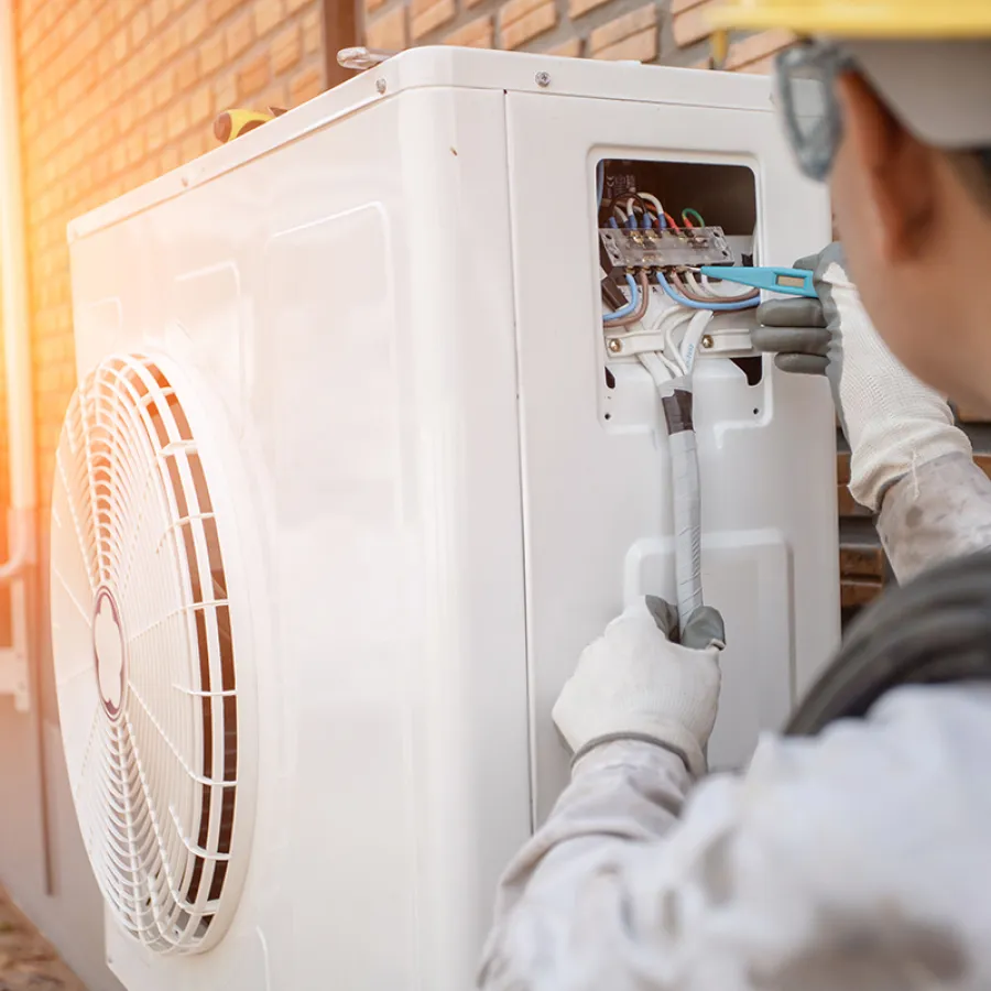Technician repairing and wiring an outdoor air conditioning unit during sunset by a brick building.