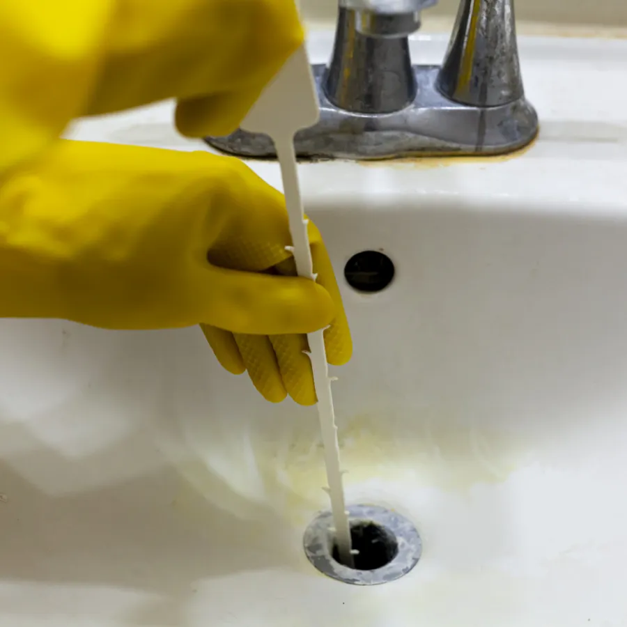 Person wearing yellow gloves unclogging a bathroom sink drain with a white tool under silver faucet.