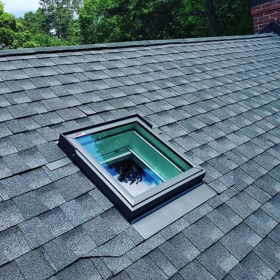 Modern black-framed skylight window installed on dark gray asphalt shingle roof with surrounding trees visible.