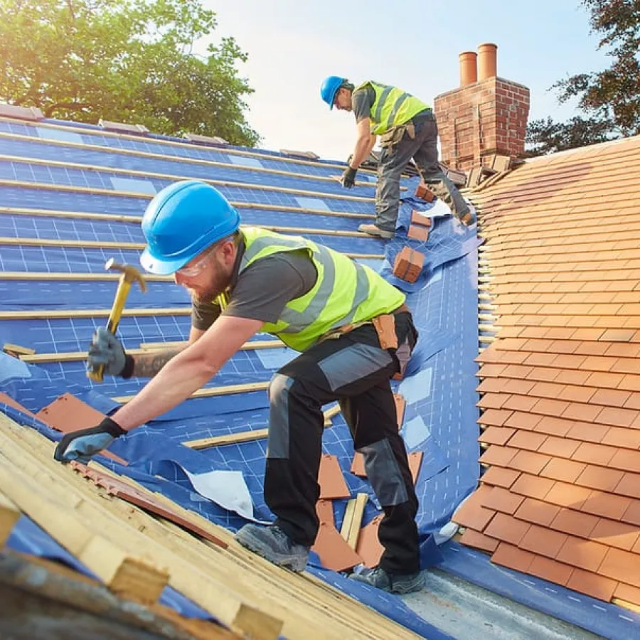Two construction workers in blue helmets installing roof tiles on a sunlit house roof during daytime.