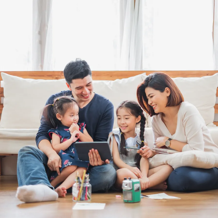 Happy family of four sitting on living room floor using tablet and smiling together.
