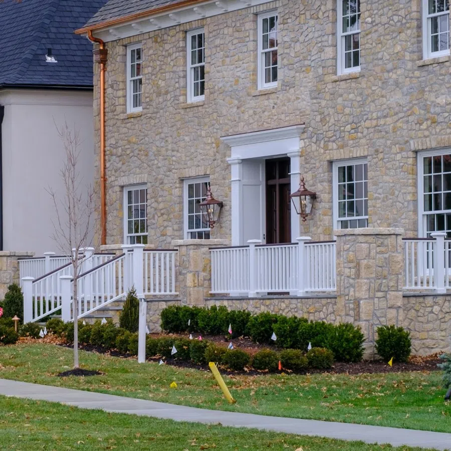 Stone house with white railing porch, copper downspouts, landscaped lawn, and large windows under a gray roof.