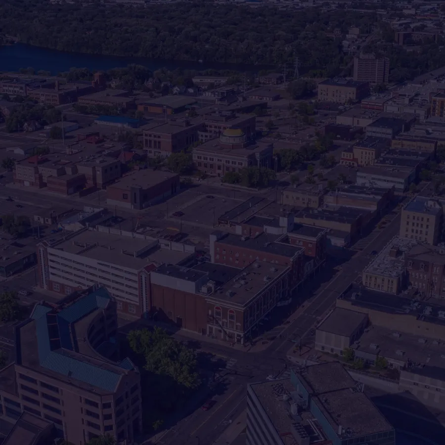 Aerial view of a mid-sized cityscape with buildings, streets, and a river bordered by dense trees in the distance.