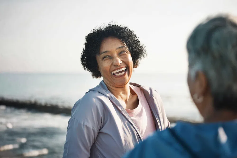 A joyful woman with curly hair laughing by the ocean, engaging with another person partially seen from behind.
