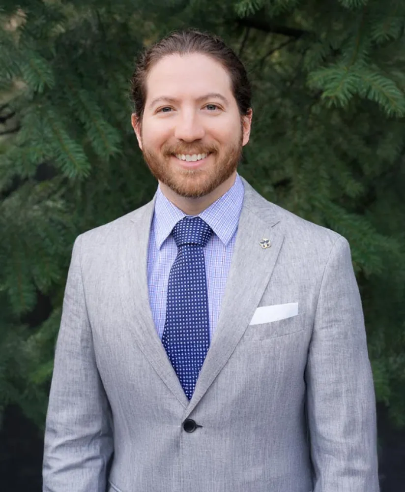 Smiling man with beard wearing a gray suit, blue shirt, and patterned tie standing in front of green foliage.