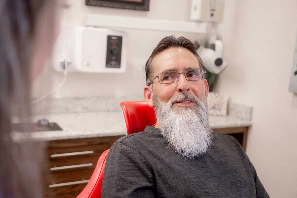 Bearded man with glasses sitting on a red dental chair talking in a dental office setting.