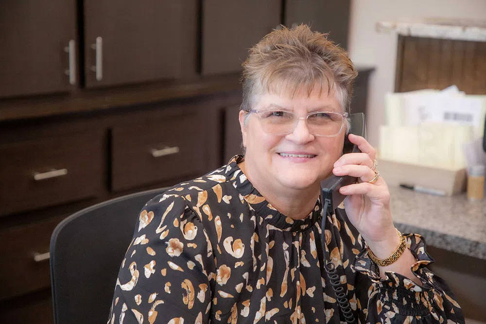 Smiling woman with glasses talking on phone in an office setting with wooden cabinets and desk.