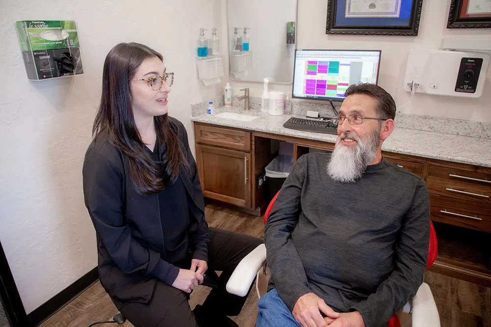 Dentist consulting with male patient in modern dental office with computer and certificates on wall.