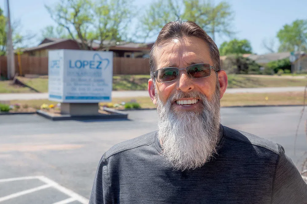 Smiling man with long gray beard and sunglasses standing outdoors near Lopez Chiropractic sign on sunny day.