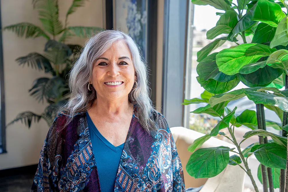 Smiling older woman with gray hair wearing a blue patterned shawl next to a large green houseplant by a window.