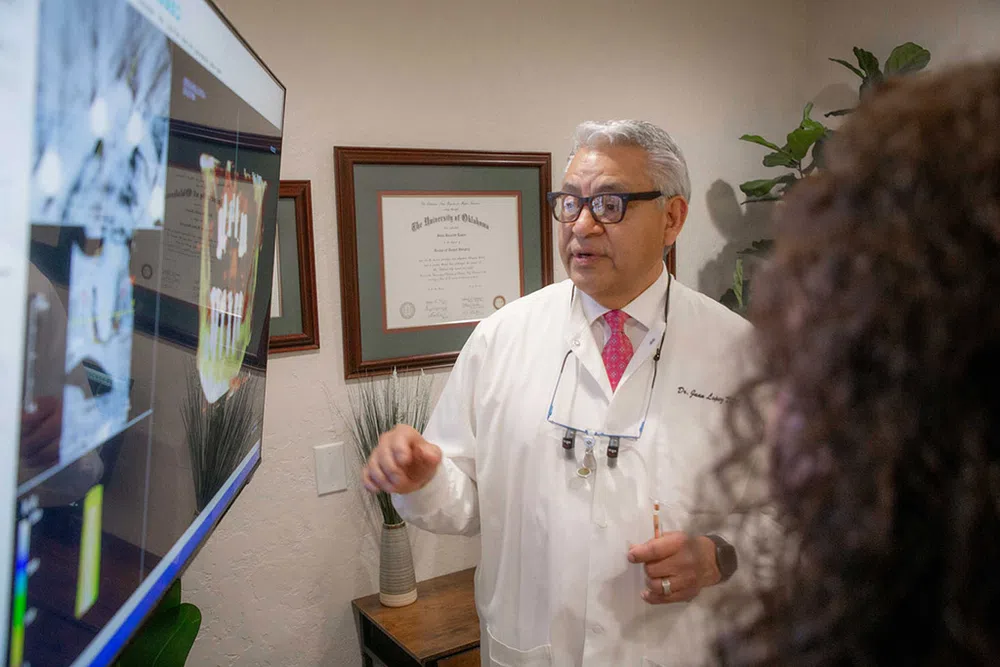 Dentist explaining dental X-rays on screen to a patient in a consultation room with framed diplomas.