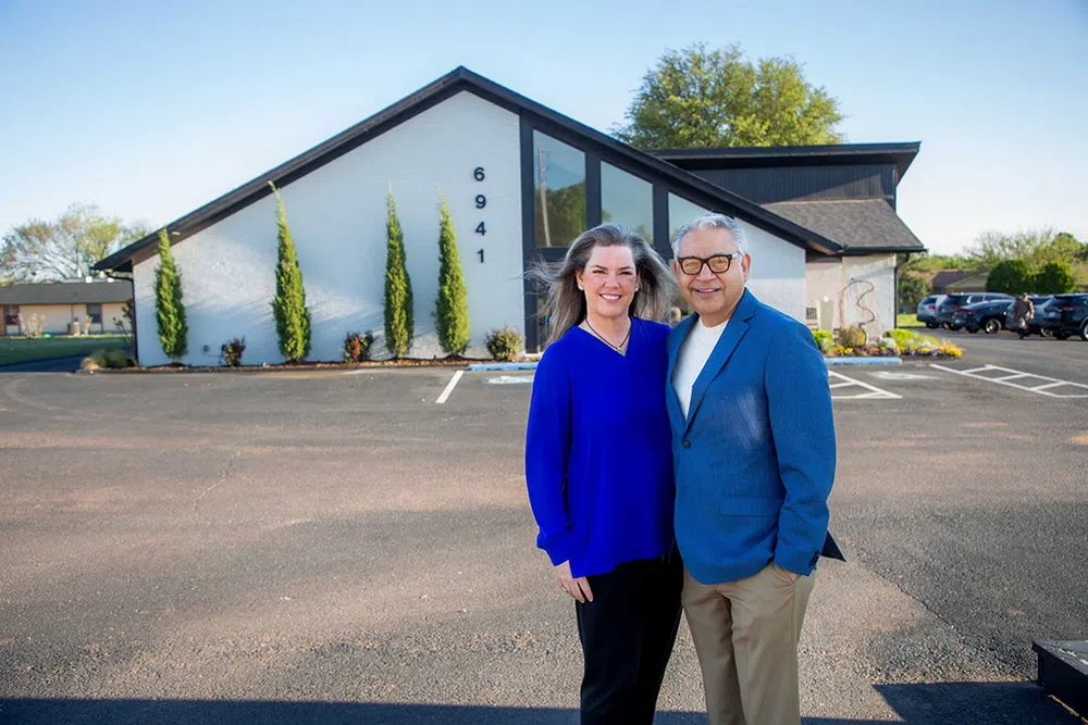 Smiling couple standing outside modern white building with address 6941 on a sunny day in parking lot.