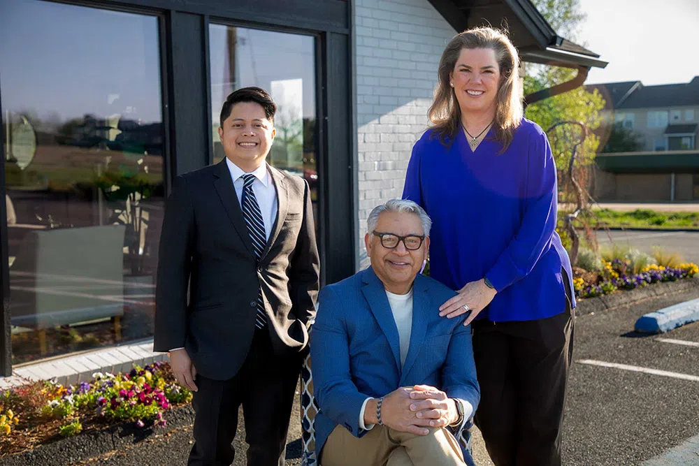 Three diverse professionals posing outside a modern office building on a sunny day, dressed in business attire.