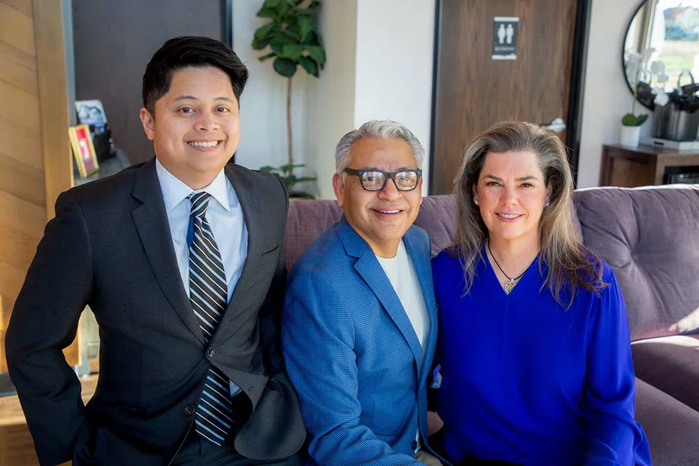 Three professionally dressed people smiling indoors on a couch with modern decor and natural light.