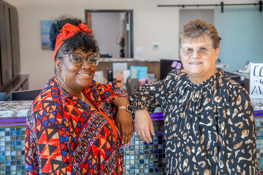 Two smiling women standing and leaning on a mosaic tiled counter in a brightly lit interior space.