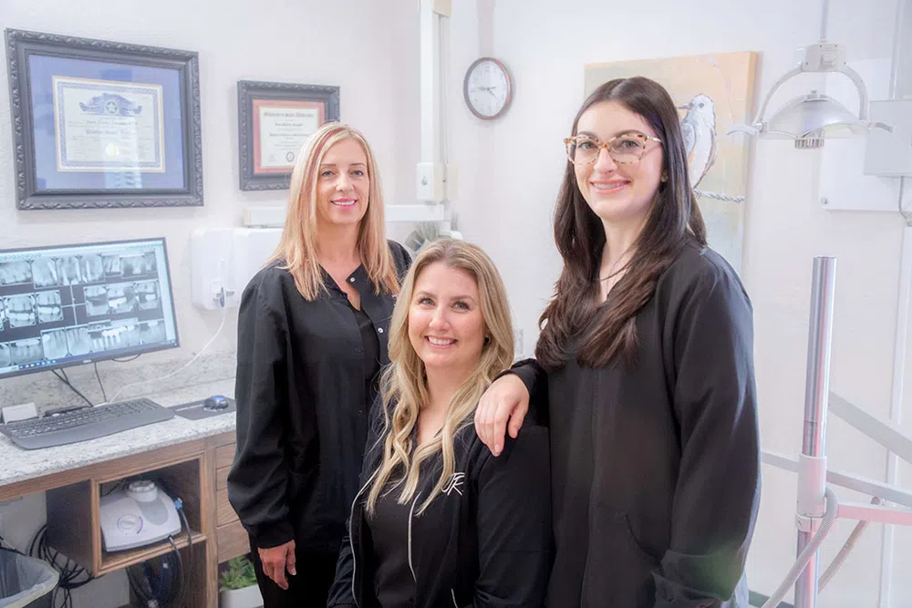 Three female dental professionals in black uniforms smiling inside a bright dental office with certificates and equipment.