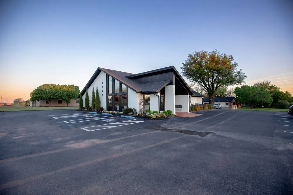 Modern building with large glass windows and landscaped greenery surrounded by spacious asphalt parking lot at sunset.