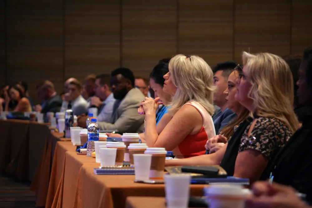 Diverse audience attentively listening at a conference table with notebooks and coffee cups.