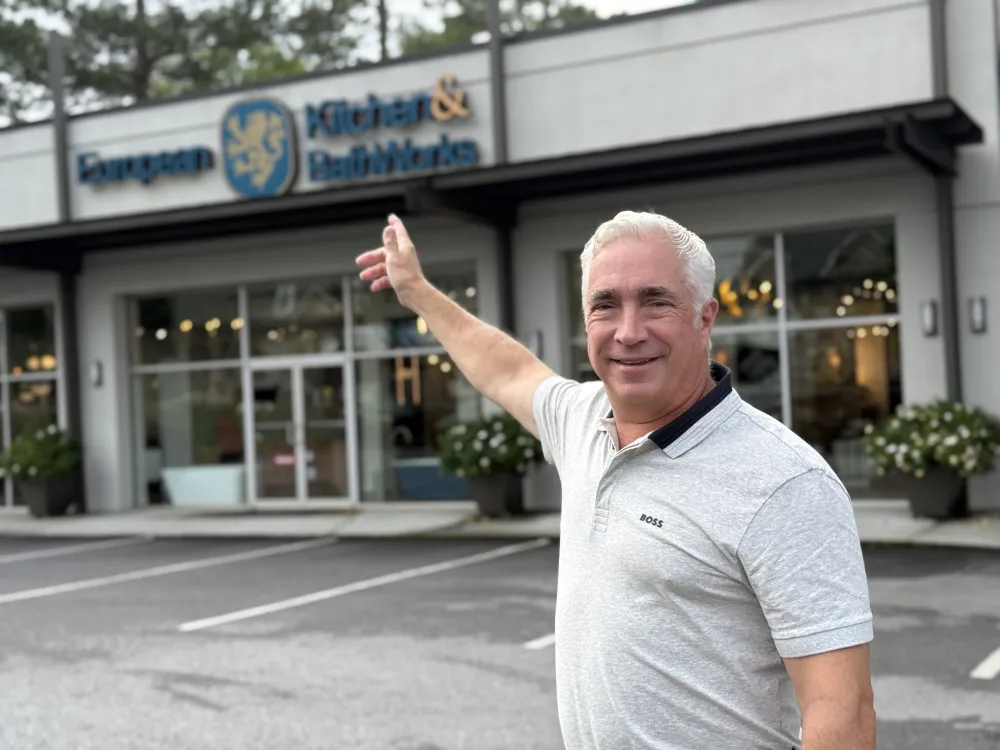 Smiling man in gray polo shirt pointing at European Kitchen & Bath Works storefront with plants outside.