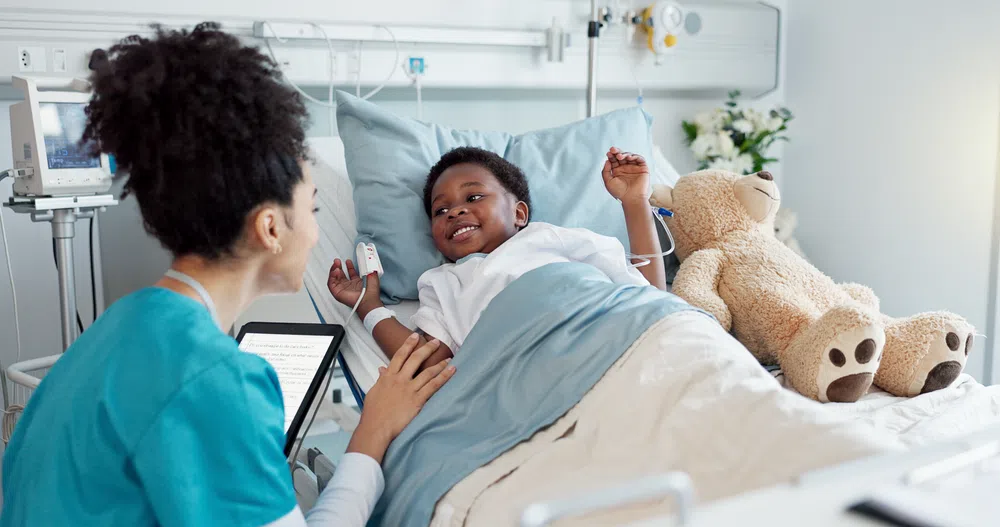 Smiling pediatric nurse interacts with happy child patient lying in hospital bed with teddy bear and medical equipment.