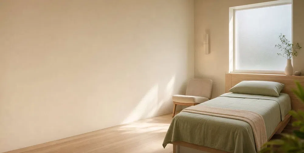 Minimalist bedroom with single bed, light green bedding, beige chair, and natural sunlight through frosted window.