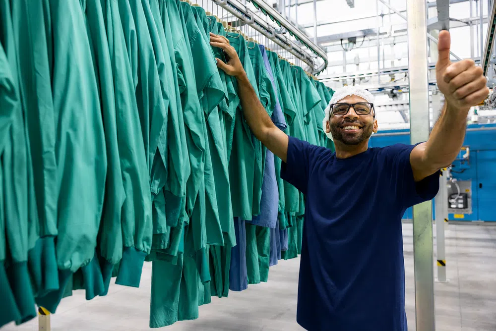 Smiling worker thumbs up next to hanging green medical scrubs in an industrial laundry facility.