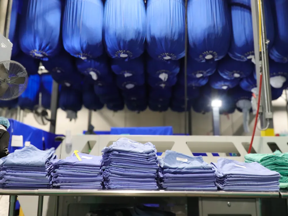 Stacks of folded blue hospital scrubs on a table with bundles of blue cloth suspended overhead in a laundry facility.