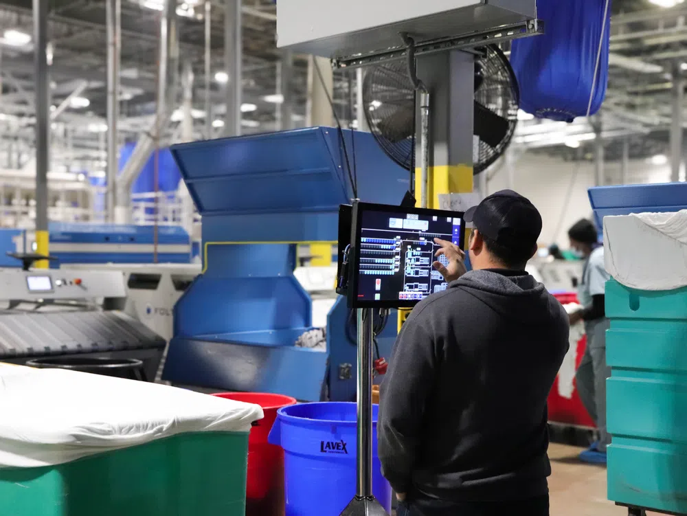Worker operating industrial touchscreen control panel in a large factory with machinery and bins around.